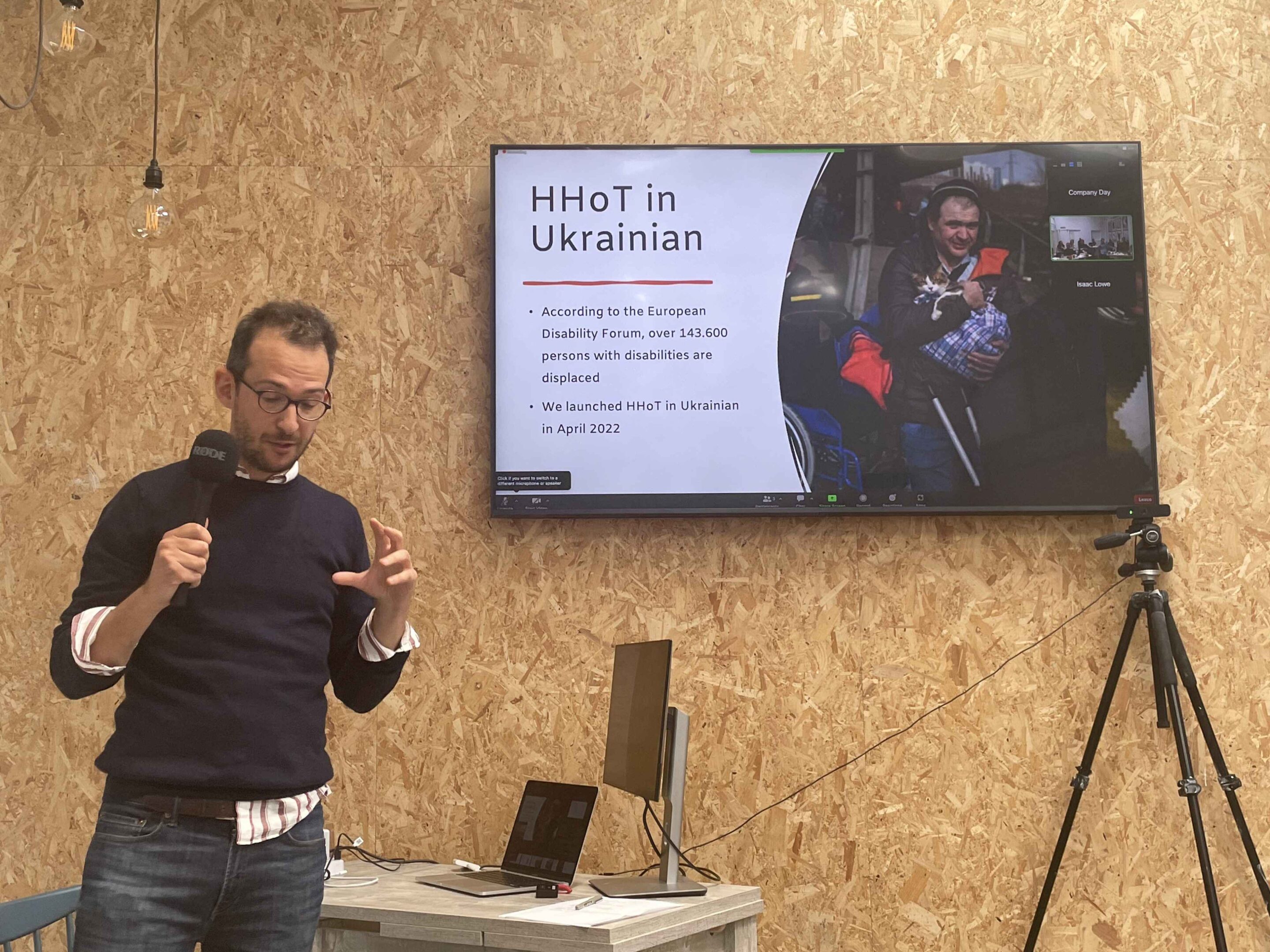 Fran is wearing a navy jumper over a white shirt, with blue jeans. He is standing in front of a presentation that says: HHoT in Ukrainian. According to the European Disability Forum, over 143,600 persons with disabilities are displaced. We launched HHoT in Ukrainian in April 2022. 