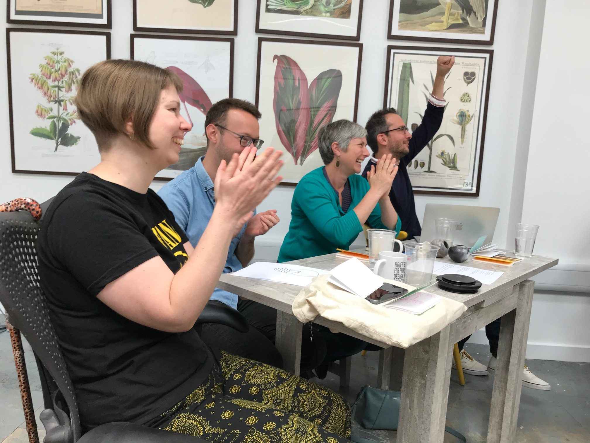 Four team members sitting in a row at a table. Nicki, Sean and Julia are clapping and laughing, Fran is punching the air. 