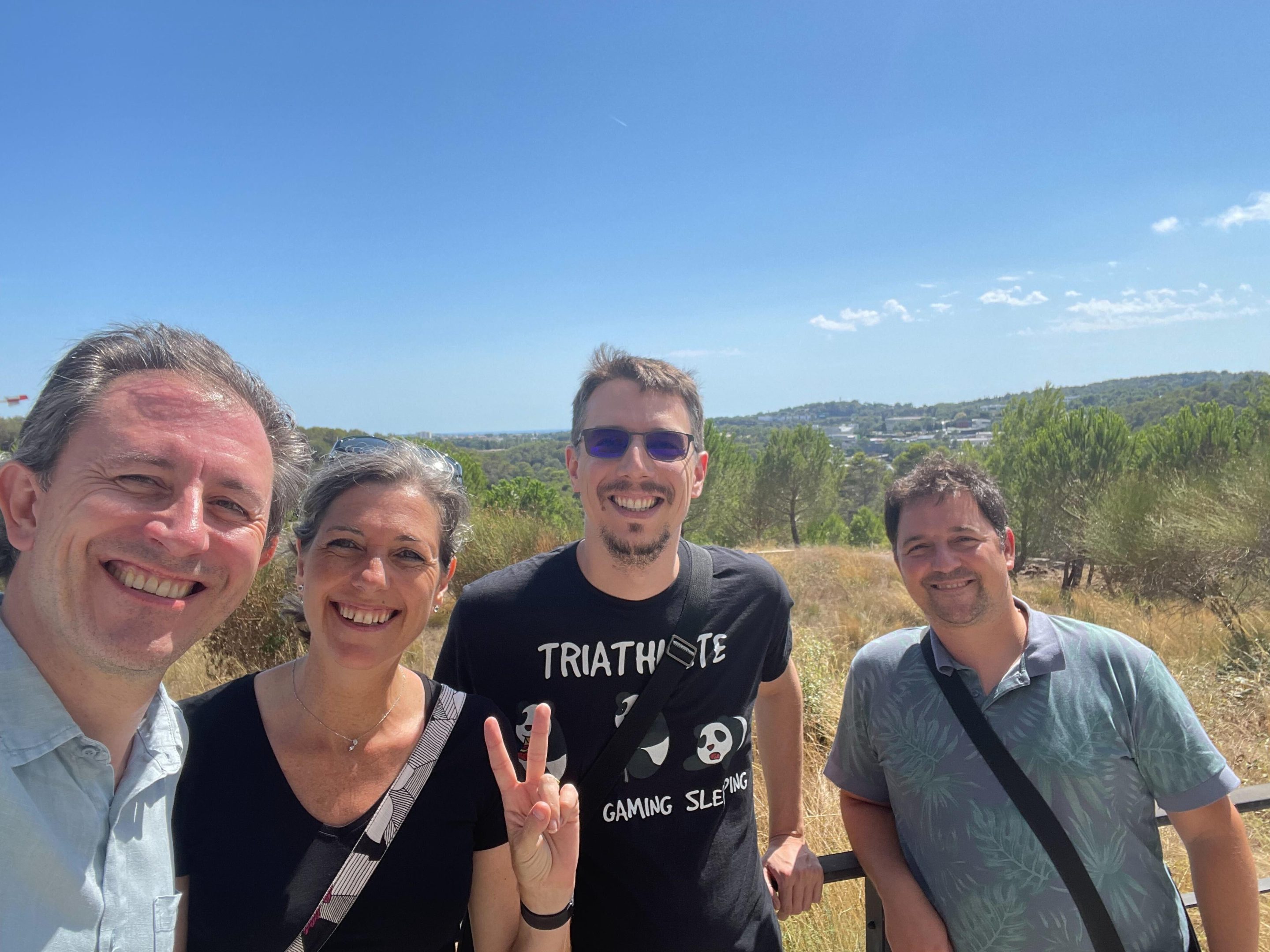 Smiling selfie of Simon with the W3C team. They are standing outside with a big blue sky and undulating landscape. Coralie is smiling and making a peace sign.