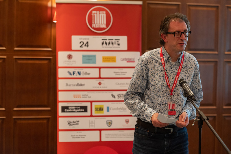 A man wearing a red lanyard is standing in front of a microphone, holding notes for a speech he is giving. A pull-up banner of film festival sponsors is behind him