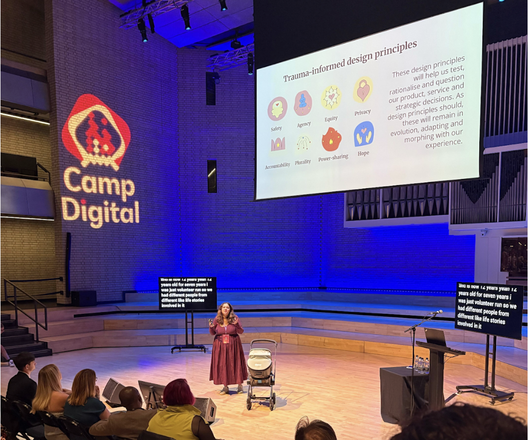 Woman stands on a stage next to a pram. The screen behind her says trauma-informed design principles.
