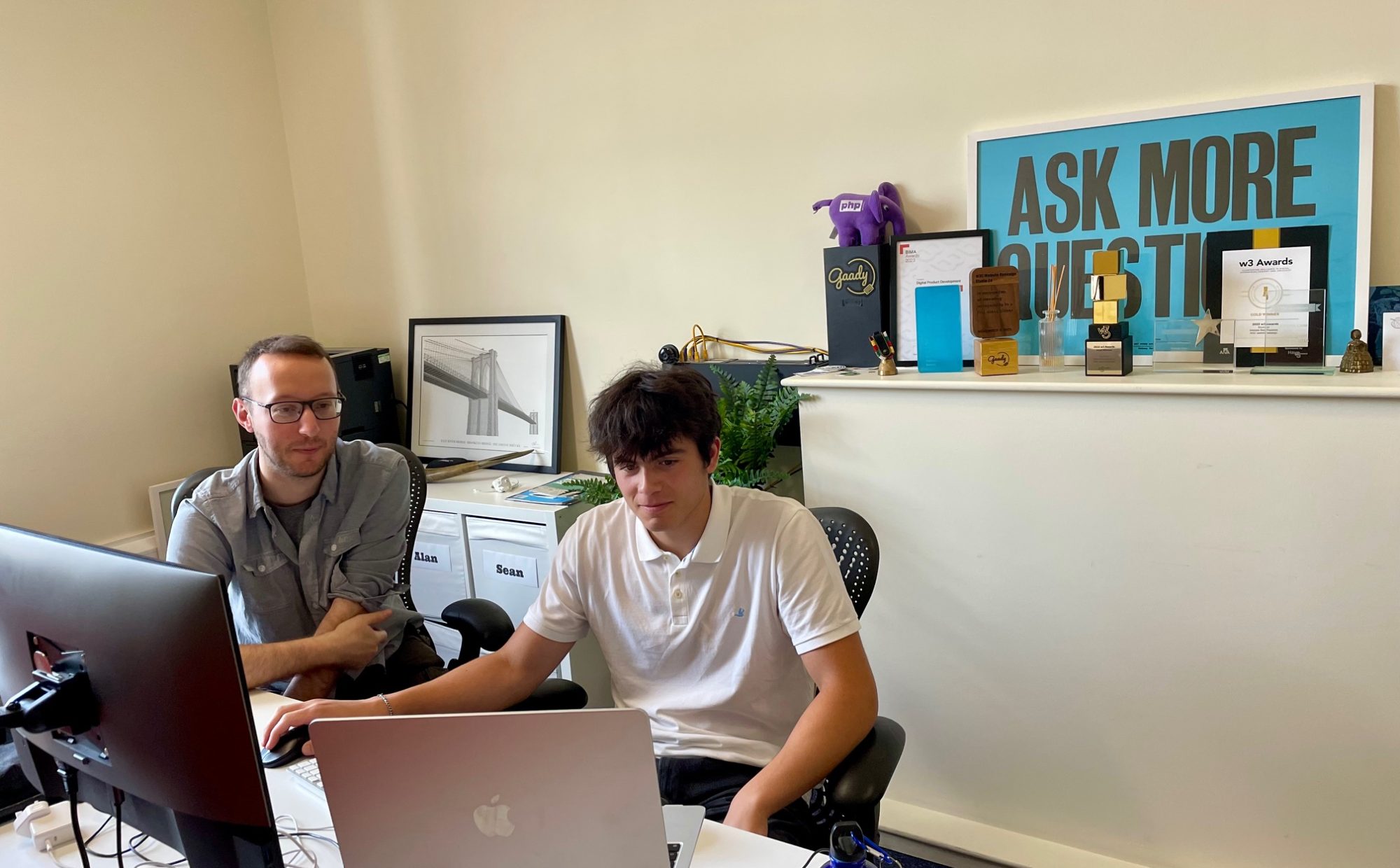 In an office, Sean and Ben are sat at a work desk in front of a laptop and monitor. Ben has his hand on a mouse. A framed poster behind them reads Ask more questions.