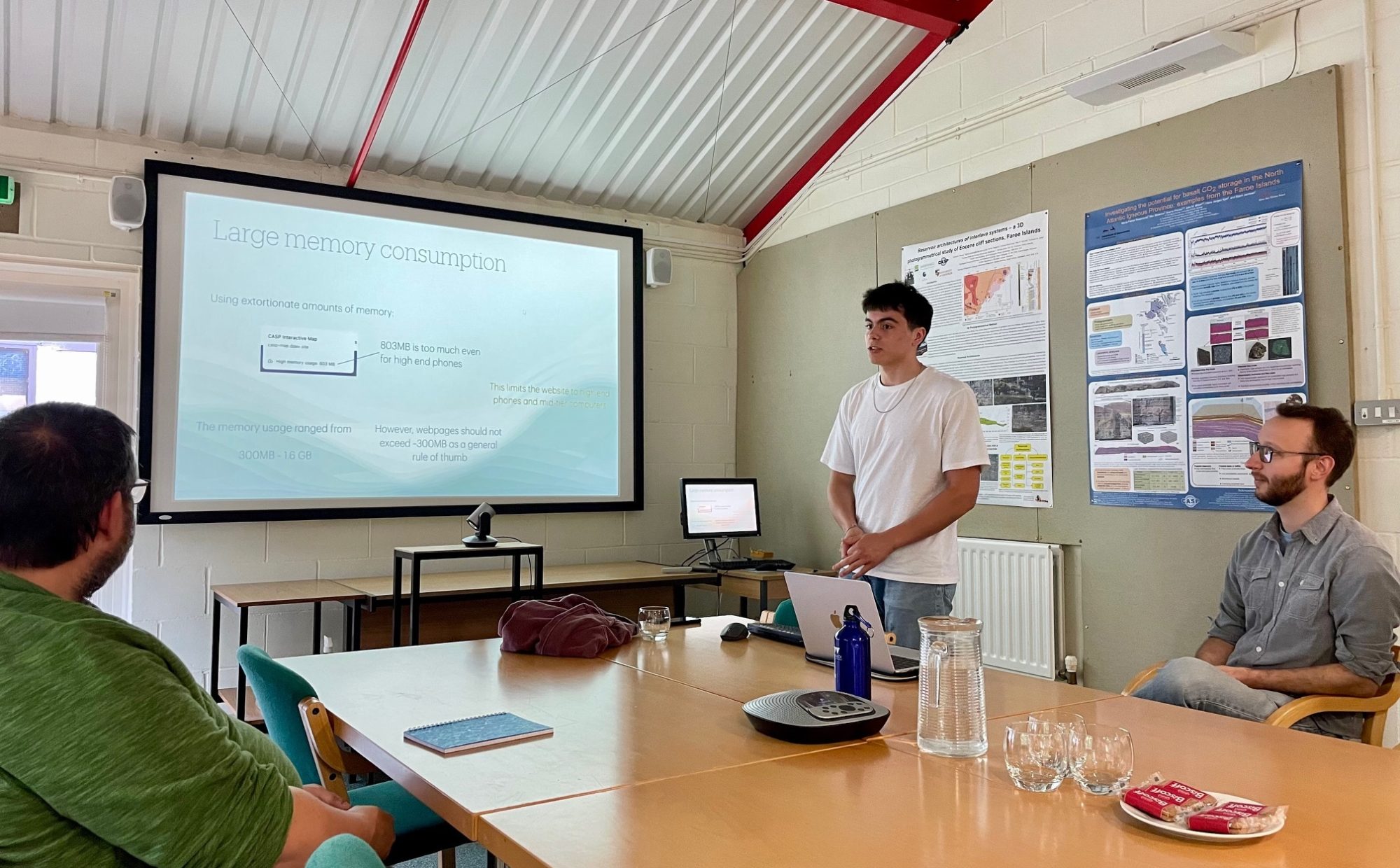 In a meeting room. Sean and Simon are sat at a meeting table looking at a presentation screen. The slide on display says Large memory consumption. Ben is standing in front of a laptop, talking to Simon.