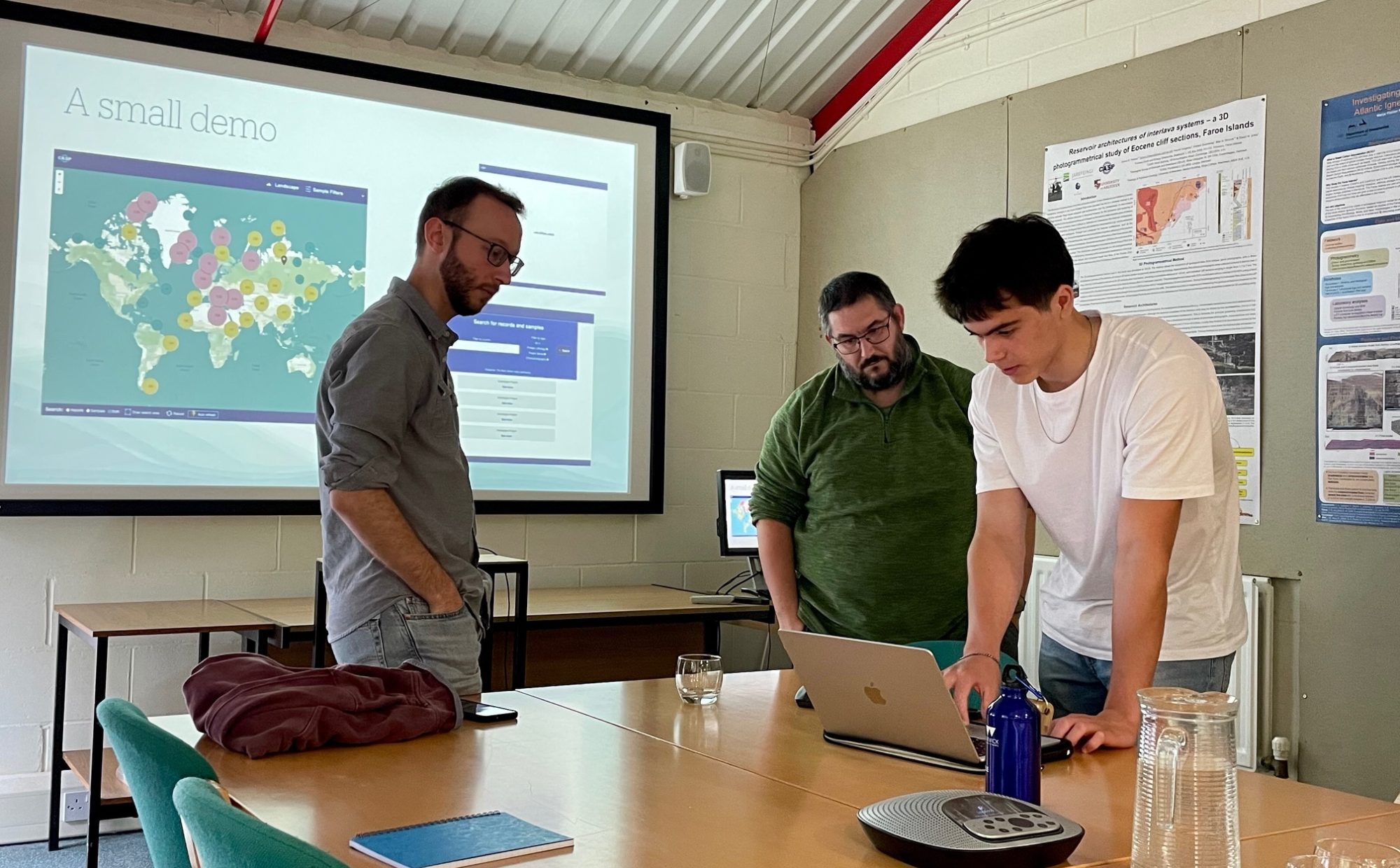 In a meeting room, Ben is showing Simon and Sean something on a laptop.