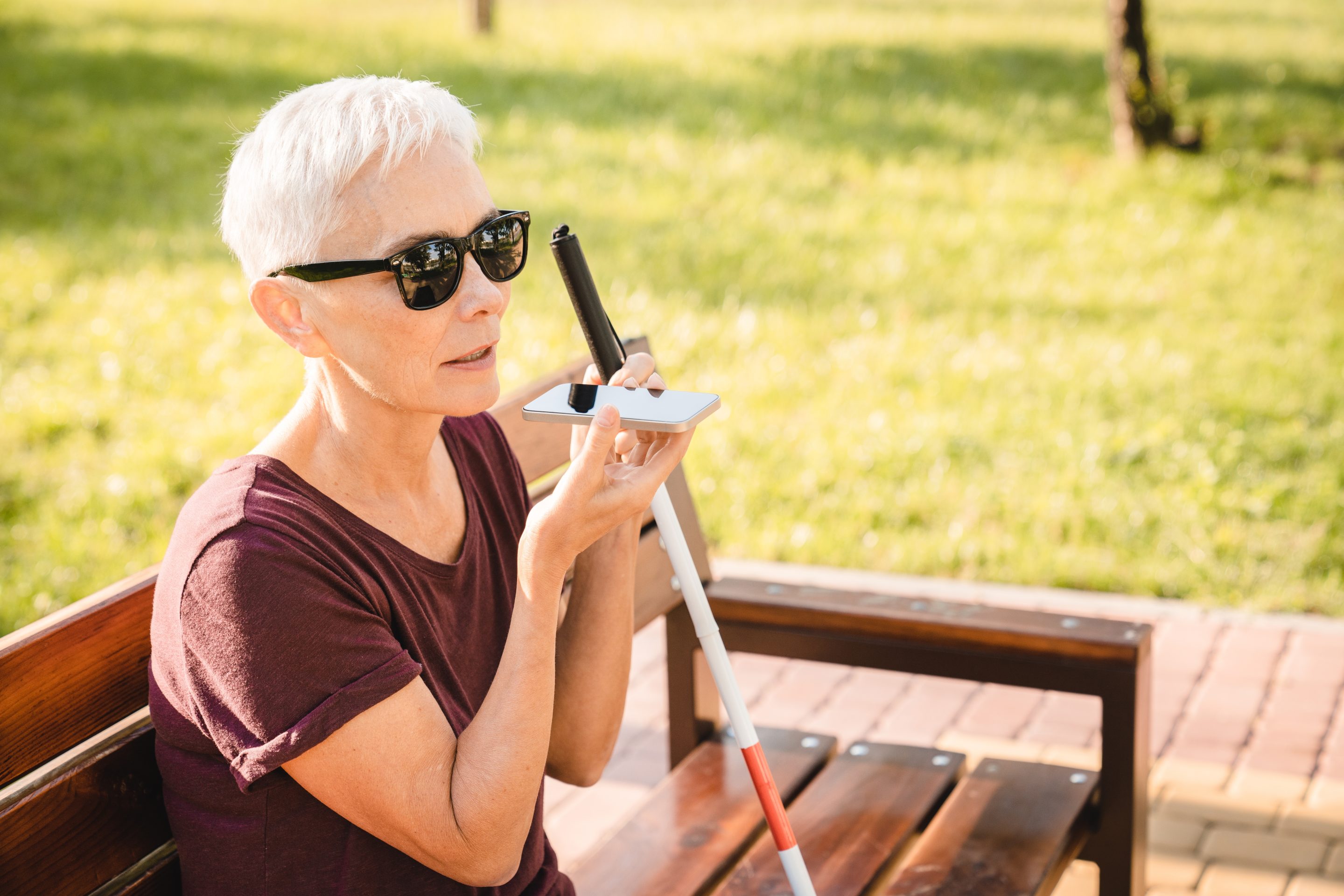 A lady with short white hair is sitting on a park bench on a sunny day. She is wearing dark glasses and is holding a white cane. She is having a conversation on her mobile phone.