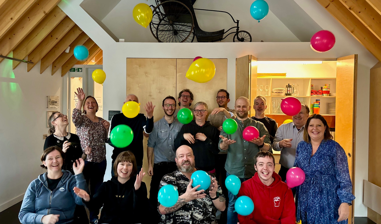 A group of smiling people standing and sitting in a large white room with wooden roof beams. Multicoloured balloons are in the air being bounced by the people.