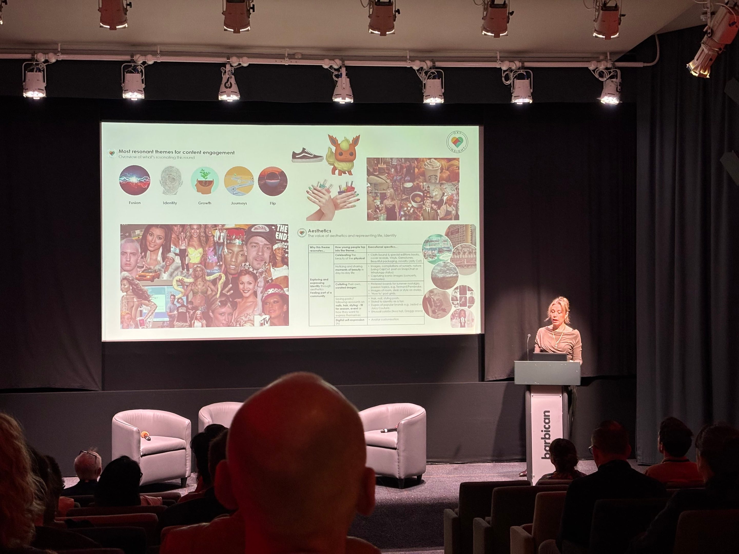 In a dark auditorium, with heavy black curtains around a small stage, a woman stands at a lectern. There is a busy slide behind her with photo montages of young people. 