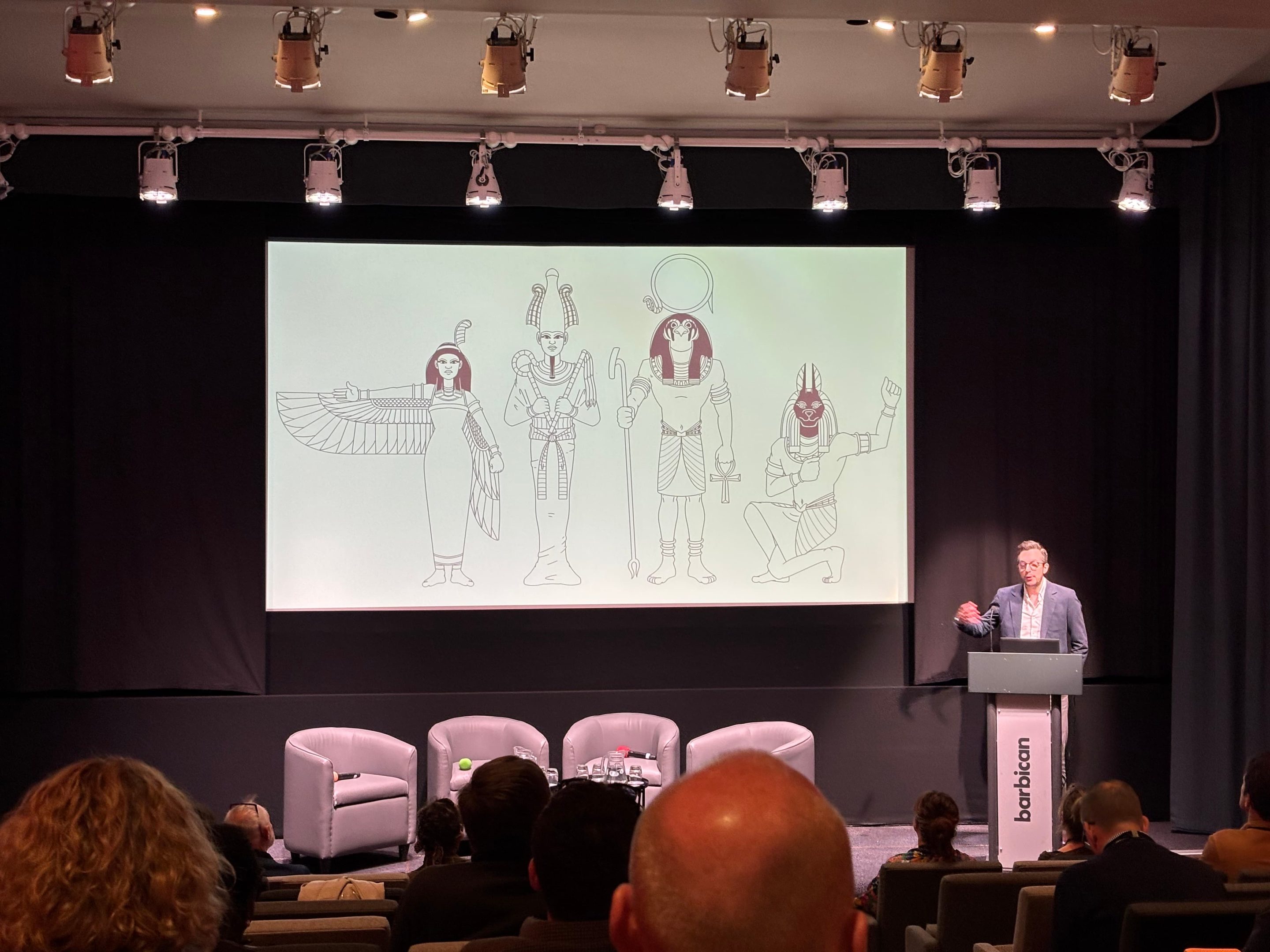 In a dark auditorium, with heavy black curtains around a small stage, a man stands at a lectern. The slide behind him has black and white line drawings of 4 Egyptian figures.
