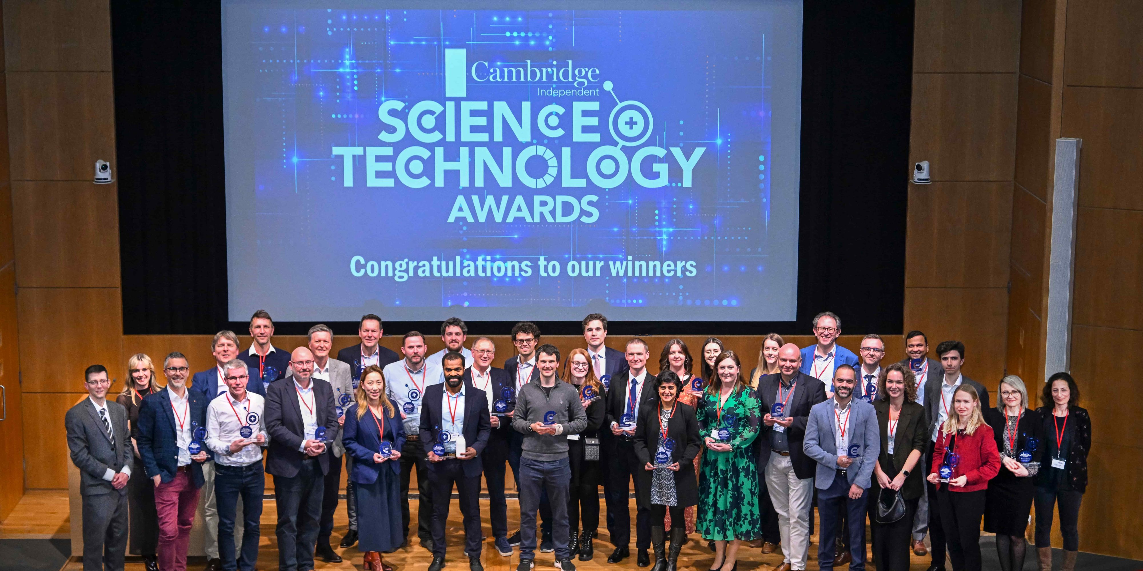 In a wood panelled auditorium, a group of about 50 people stand on a stage. The screen behind them says Cambridge and Sience Technology Awards, congratualtions to our winners.