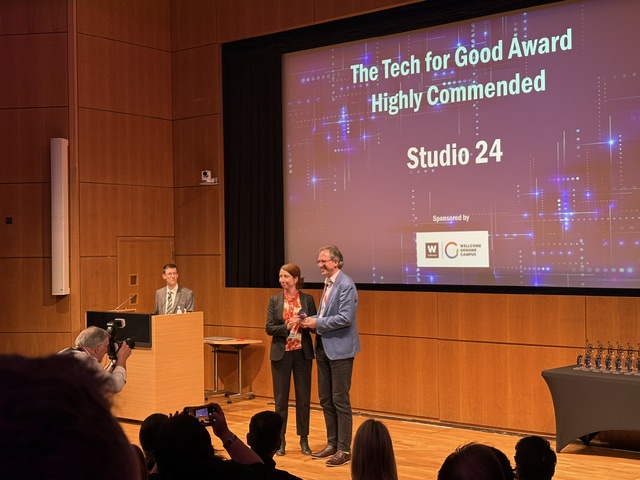 In a wood panelled auditorium, a woman has presented a man with an award and they are posing for a picture. The screen behind them reads the tech for good award, highly commended, studio 24.