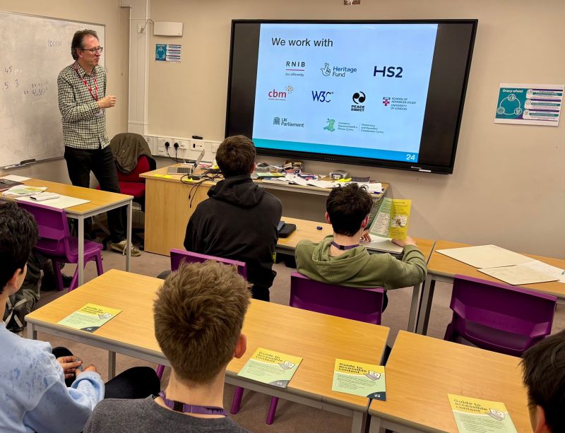 In a classroom, with desks in theatre style, a man is presenting to a room of older teenagers.