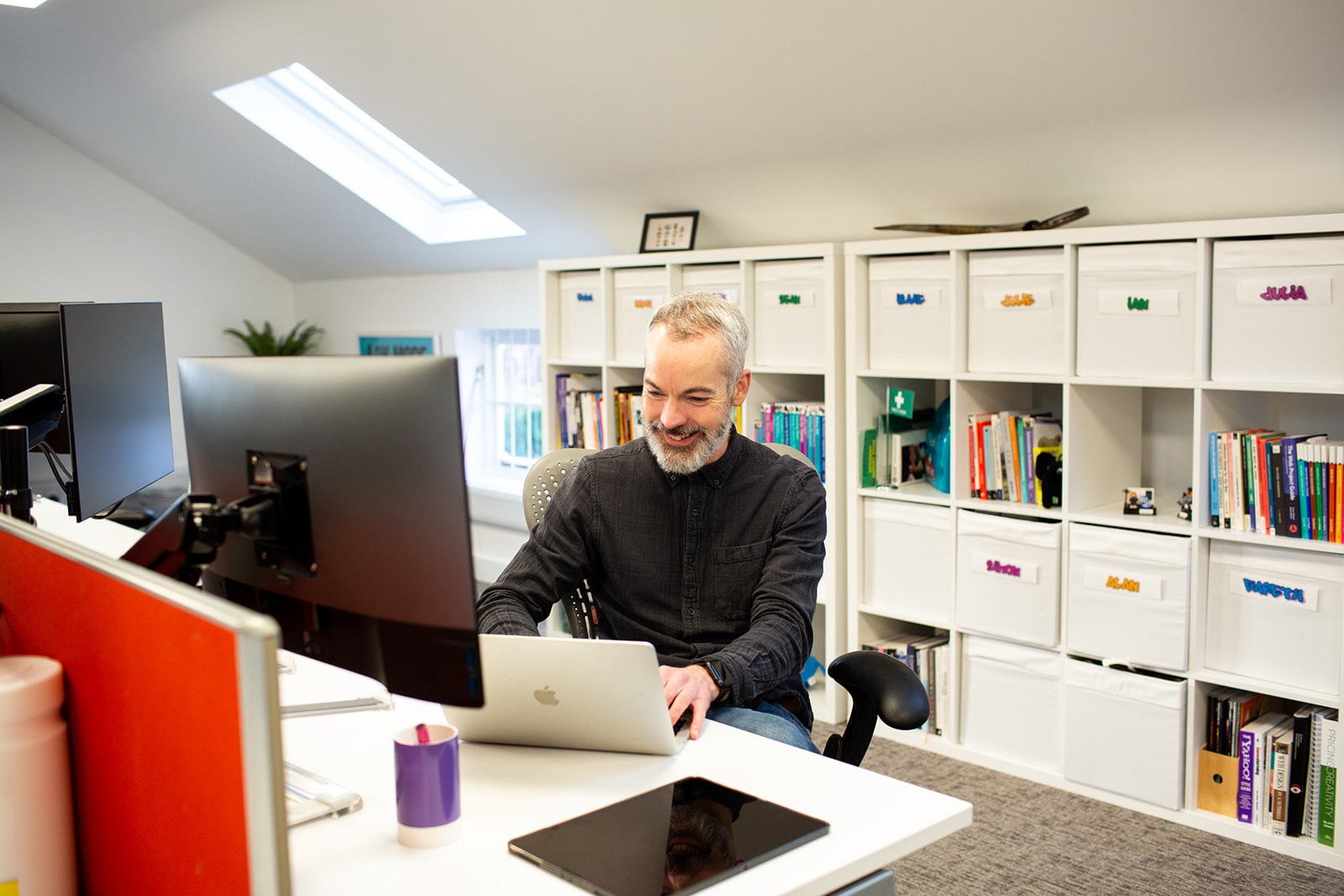 In a modern office with white walls, a man sits at a desk working on a laptop. He is smiling.
