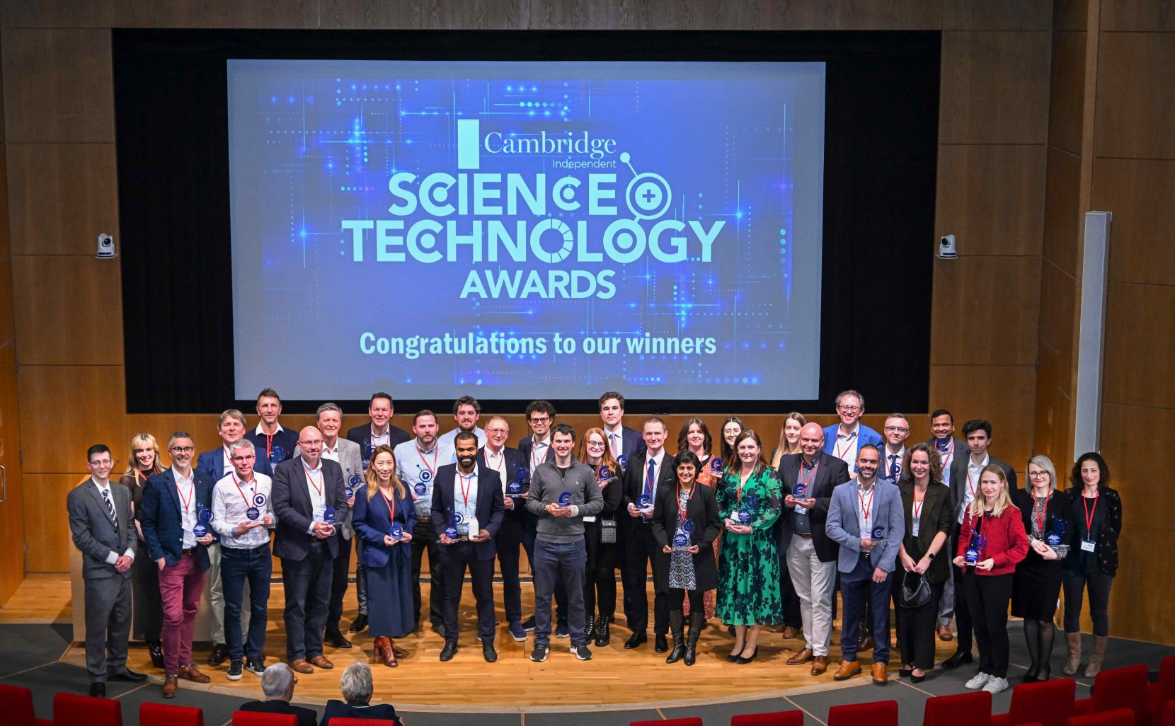 In a wood panelled auditorium, a group of about 50 people stand on a stage. The screen behind them says Cambridge and Sience Technology Awards, congratualtions to our winners.