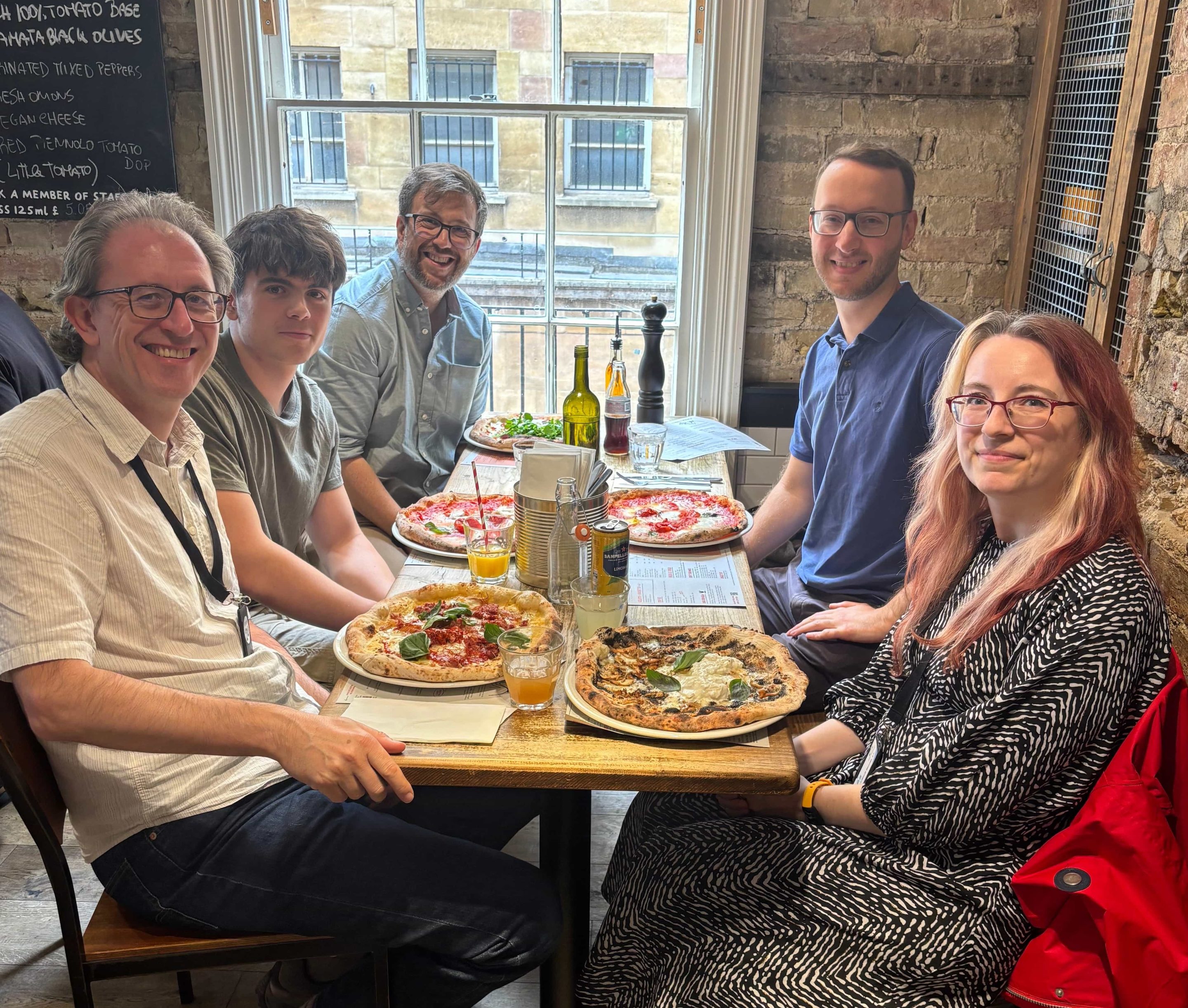 In a restaurant, four men and a woman site at a table. Pizzas have just been delivered. 