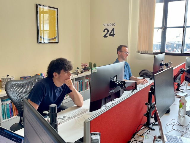 In an office, two men are sat at their desks. One is typing, the other has his chin resting on his hand. 