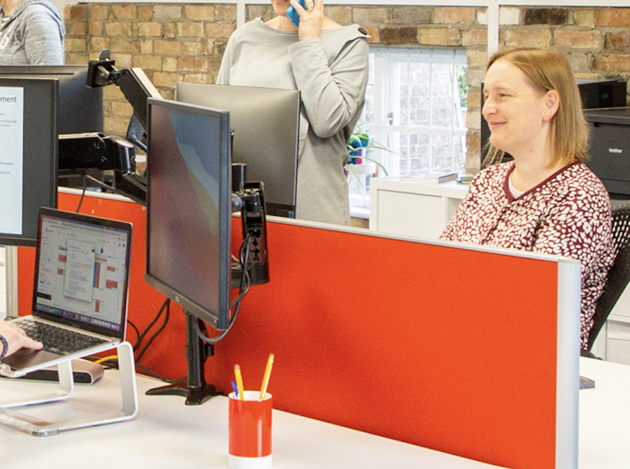 In a busy office, with lots of monitors, a woman sits behind a red desk divider. She is looking at a monitor and smiling. 